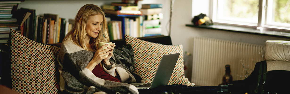 A happy woman sitting on a couch with a blanket around her drinking coffee while working on her laptop.