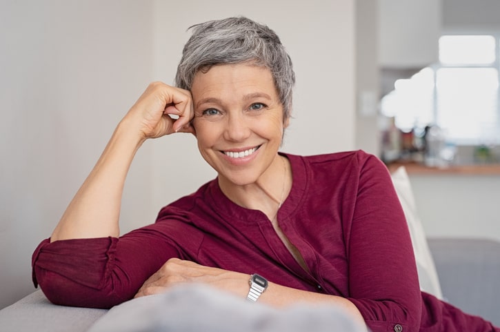 A portrait of a mature woman with salt and pepper hair smiling sitting on a couch, with a burgundy long sleeve.