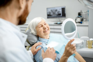 Happy elderly woman enjoying her beautiful toothy smile looking to the mirror at her new dentures in the dental office.