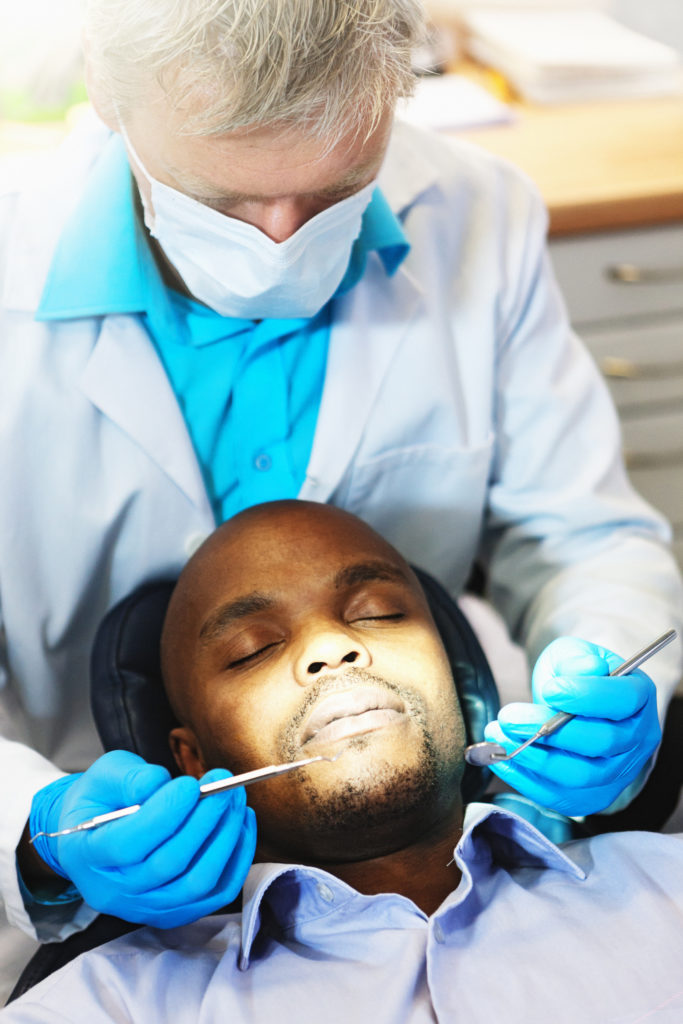 A patient lies in the dentists's chair, eyes and mouth closed, as he waits for the dentist standing behind him to begin his examination.