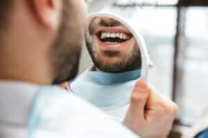 Man smiling and looking at teeth in mirror after dental crown lengthening procedure.
