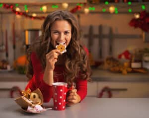 Woman during Christmas time biting into a cookie and holding a mug of hot chocolate.
